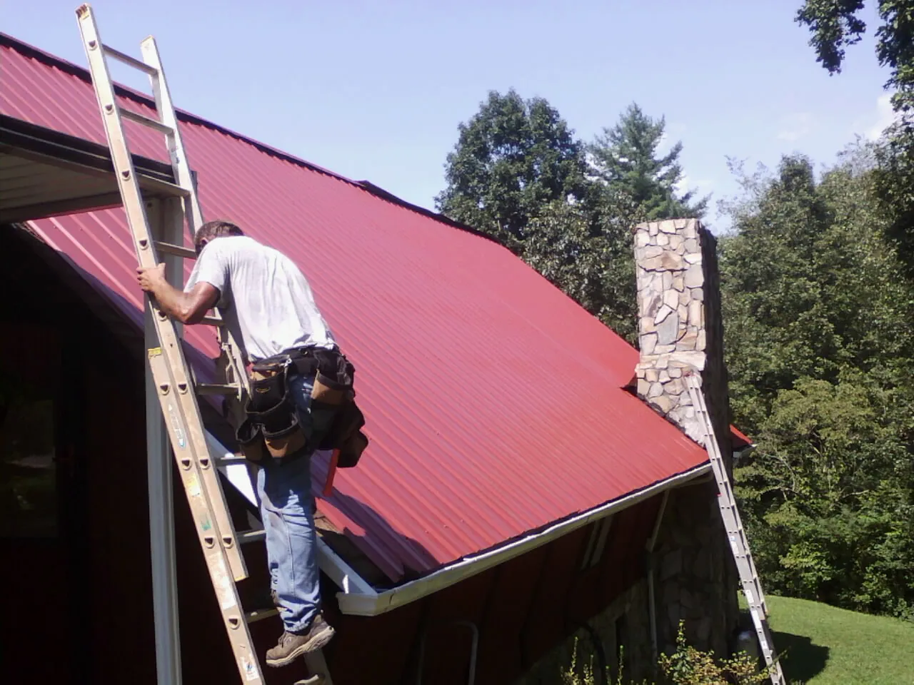 Roofer on a ladder beside a red metal roof and stone chimney — Soffit & Fascia Repair by Roofing Brothers in Middle Island