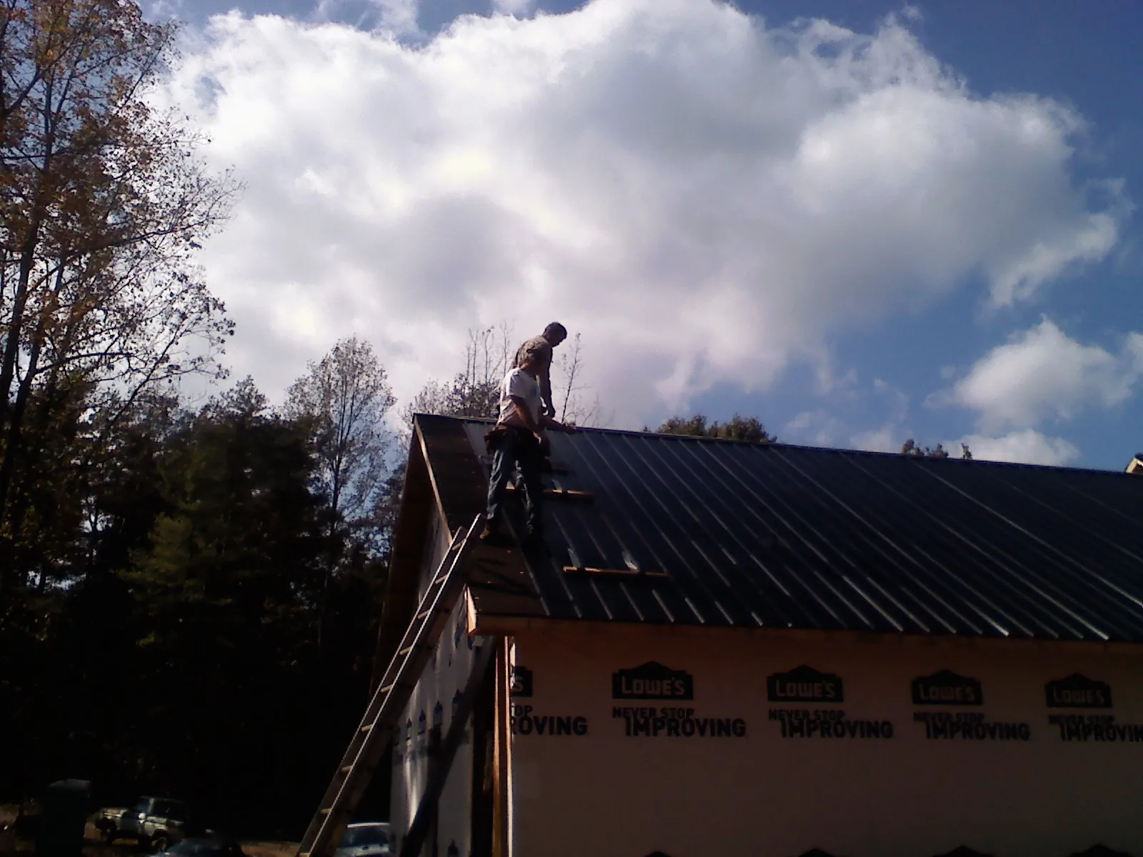 Roofer fastening dark metal panels high against a cloudy sky, from a Roof Inspection job in Middle Island